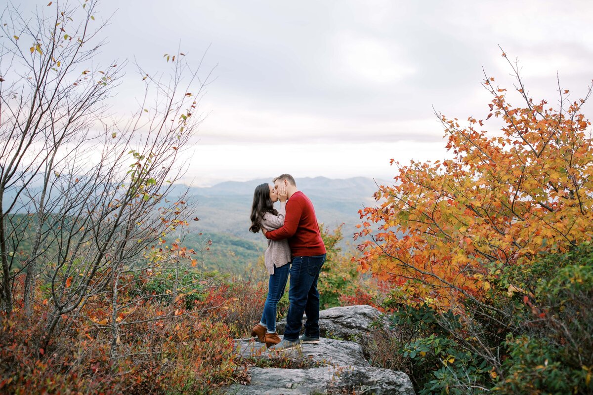 Young man in a red shirt holding a young woman with brown hair closely and giving her a kiss with the view of the Appalachian Mountains behind them in the fall
