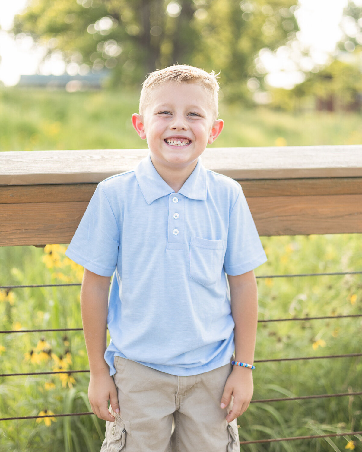 Young boy with light brown hair standing on a bridge in a wildlife sanctuary wearing a partial button up light blue shirt, khaki shorts, and smiling at the camera