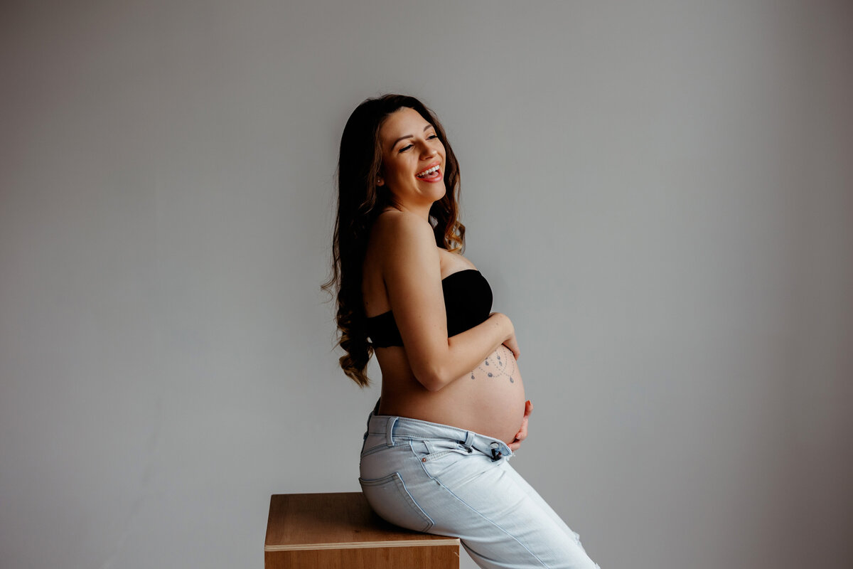 pregnant mom holds belly while wearing black bra and jeans while sitting on brown posing boxes