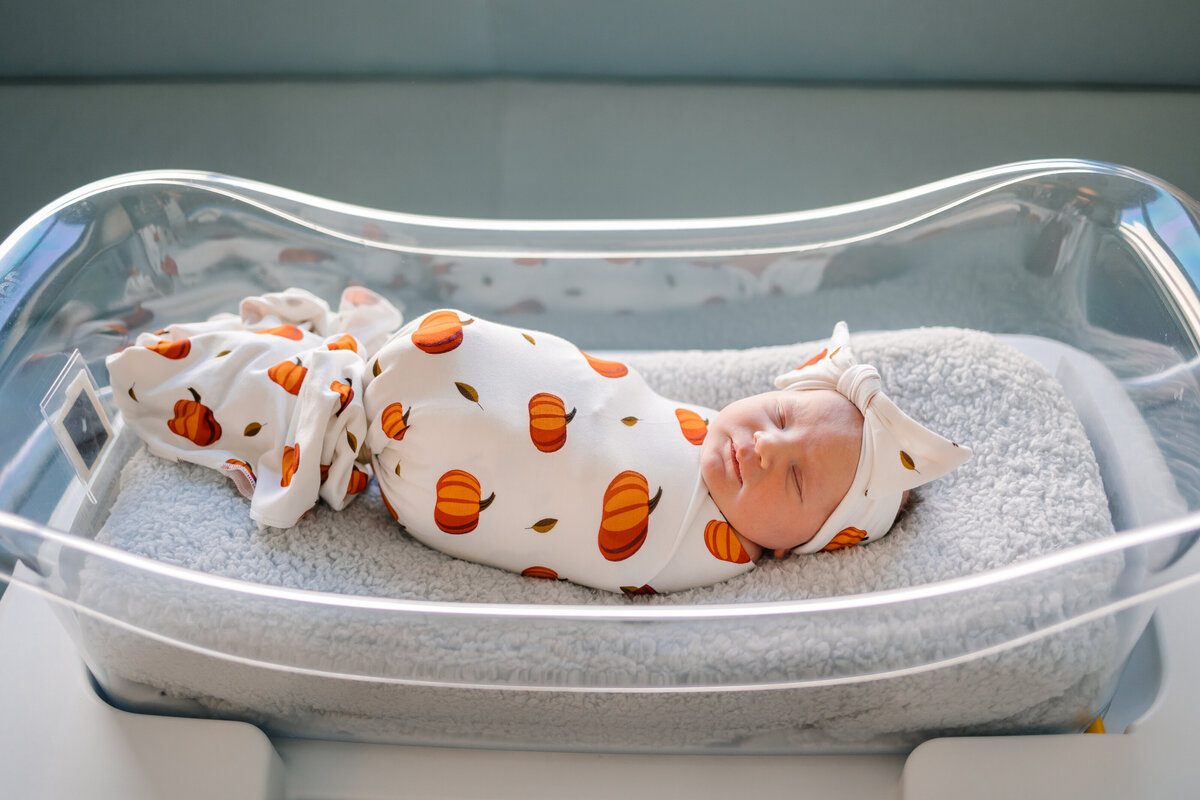 Newborn baby girl sleeping in a hospital crib, covered with an orange pumpkin blanket and wearing a white headband.