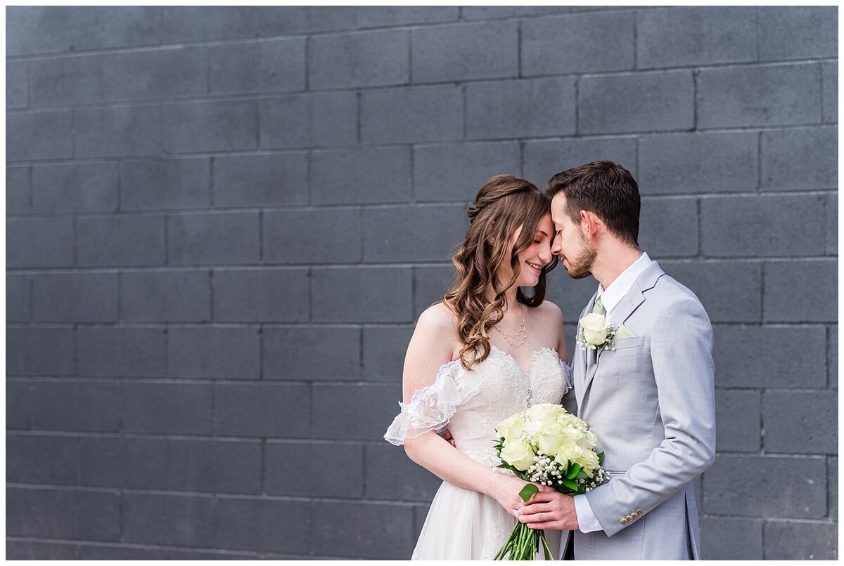 Bride and groom stand forehead to forehead while hold the bride's white rose bouquet between them while standing in front of a black wall outside of the venue skylight in downtown Denver