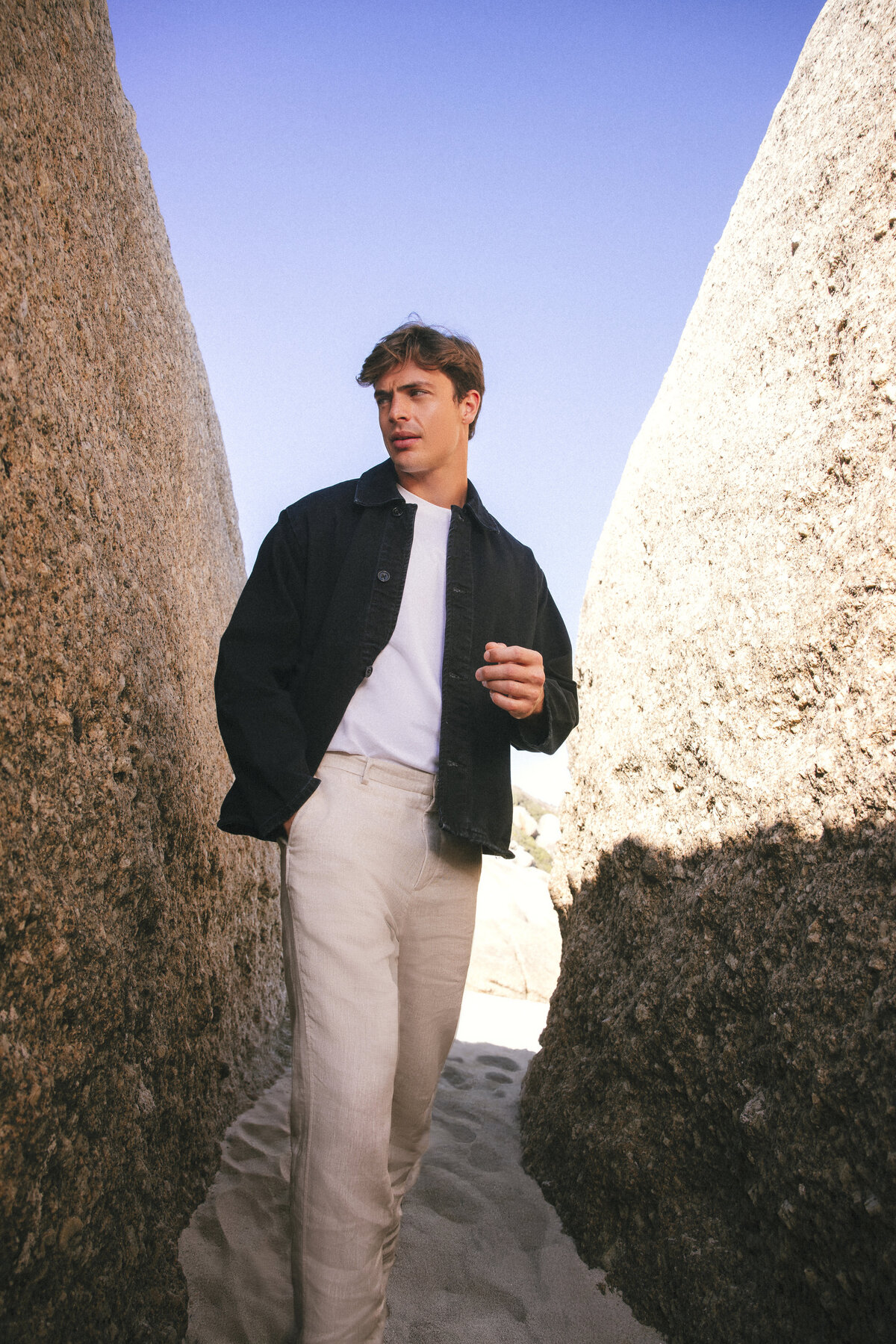 Male model leaning against a large rock in Cape Town, wearing a black denim jacket, white T-shirt, and beige trousers, with soft sunlight casting shadows on the sand.