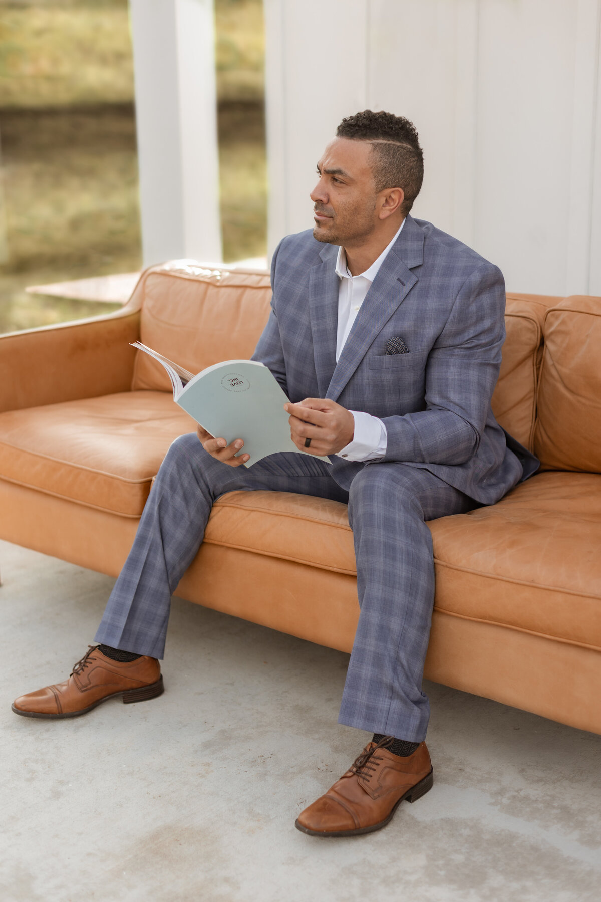 Checkered Suit, Brown Suit well build vaguely middle eastern man reading an empty book on a brown leather couch.