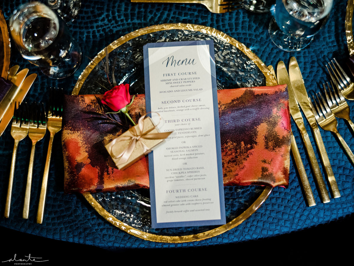 place setting with red patterned napkin, menu card, pink spray rose, on navy blue linen
