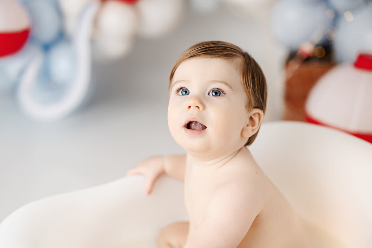 A baby with short brown hair and light skin sits in a white bathtub, looking upwards with a curious expression. Captured by a portrait photographer specializing in family photography, the softly blurred blue and red background adds to the playful atmosphere of this moment in Bloomington, Illinois.
