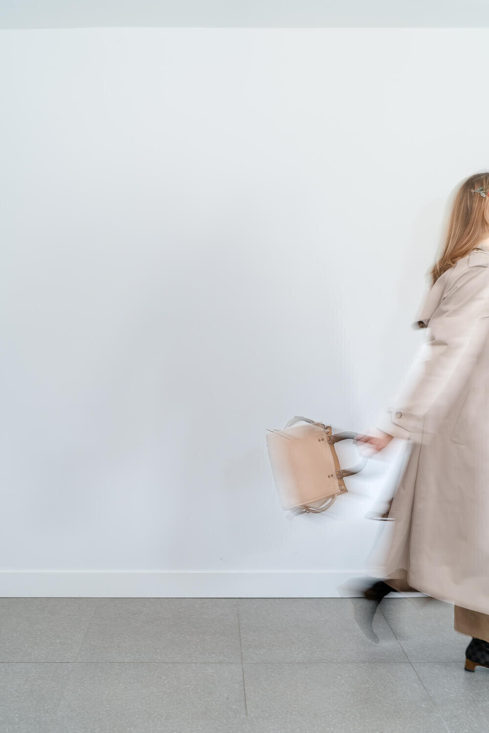 Motion-blurred woman in beige trench coat carrying structured tote bag against white wall.