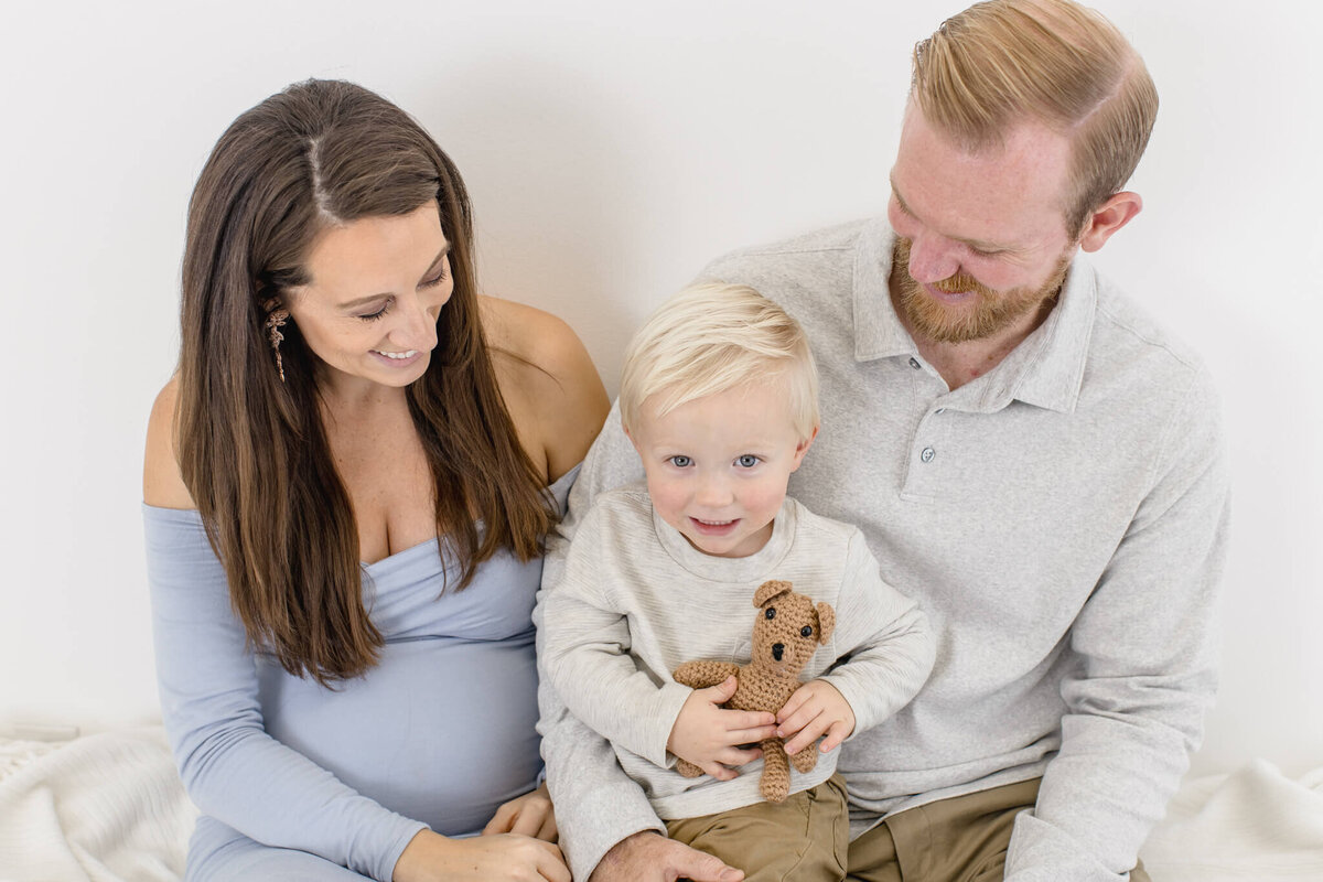 A pregnant woman, a man, and a young child sitting together on a white background. The child is holding a small brown teddy bear. The woman is wearing a blue dress, and the man is in a light gray top. They are all smiling and looking at the child.