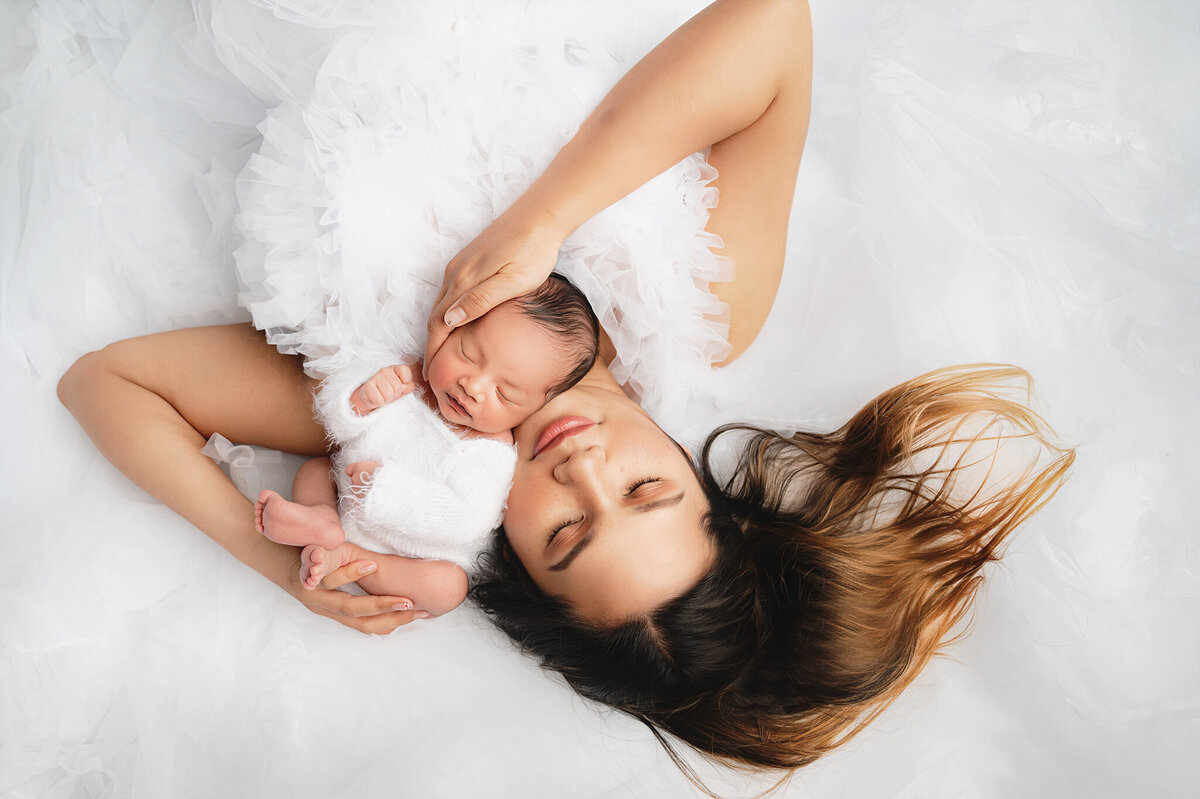 New mom wearing a white gown holding her newborn boy laying down with eyes closed, in studio by Great Toronto Newborn Photographer, Tamara Danielle.