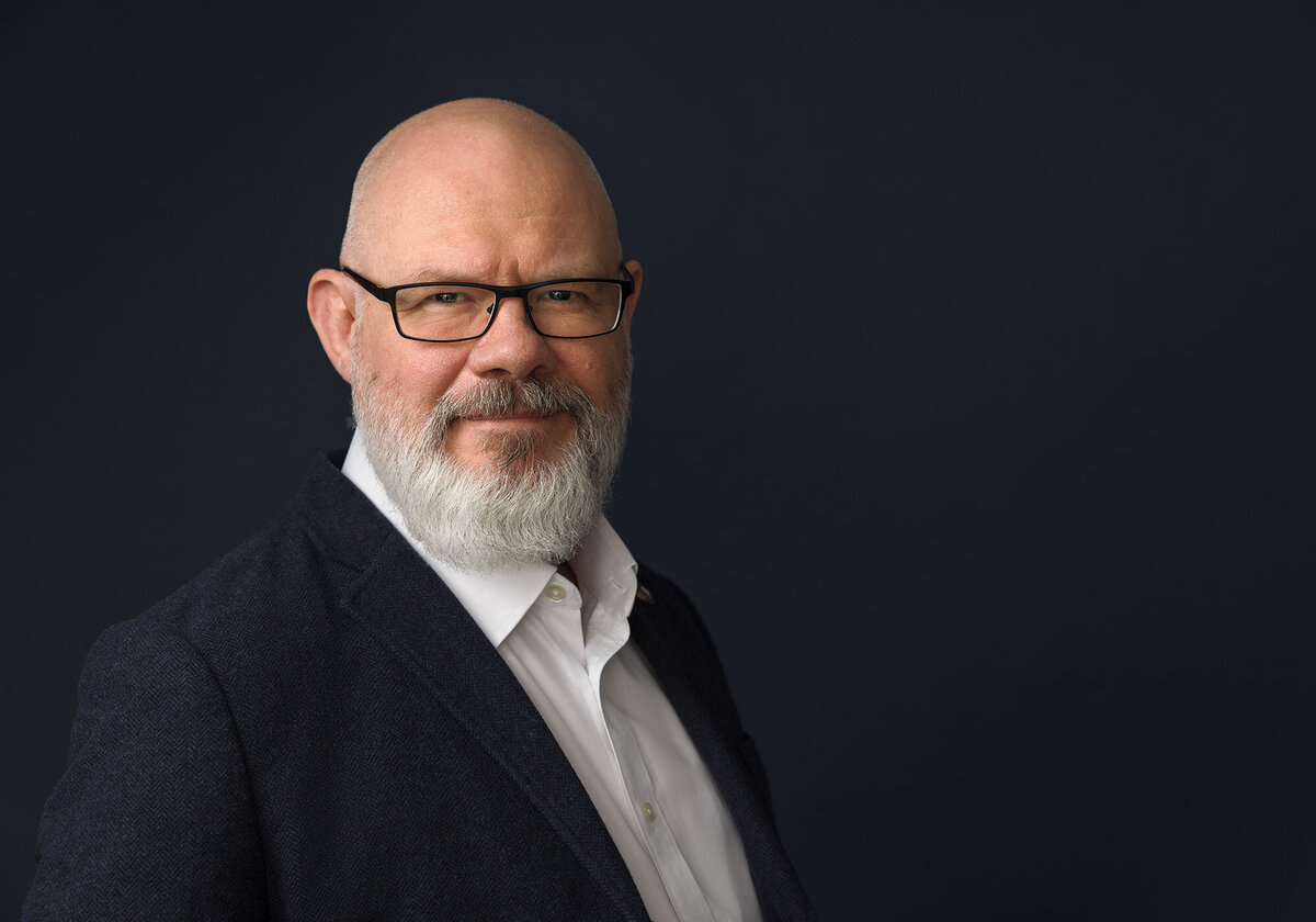 Headshot of a man with a bald head and beard wearing a navy suit. Taken in studio on a blue background by Norwich headshot photographer Claire Howes.