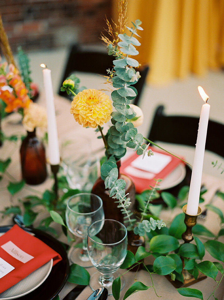 Flowers in amber glass set on a table with green garland, white plates, and lighted candlesticks.