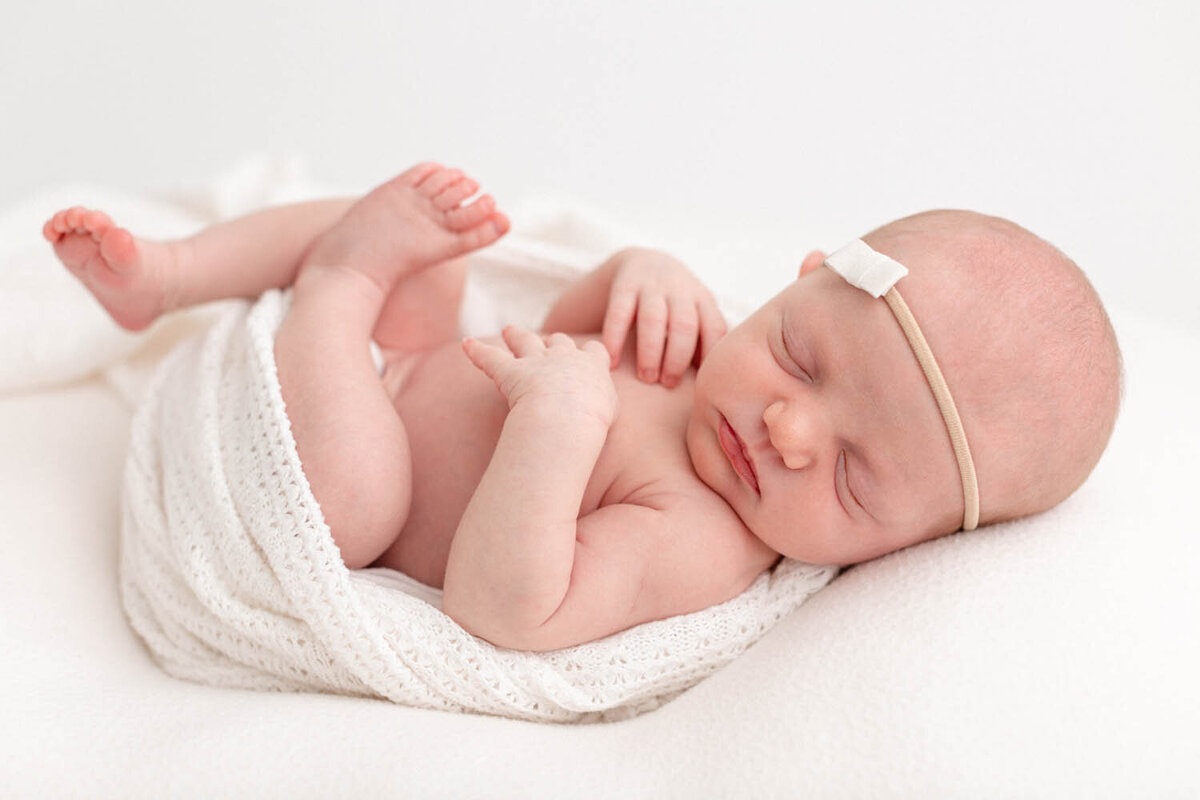 baby is sleeping peacefully wrapped in white with a tiny minimalist bow on her head.