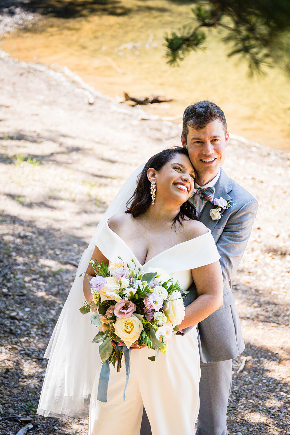 A bride stands in front of the groom and leans onto him, placing her head on his chest and holding her bouquet, while the groom looks up and smiles. The pair eloped on Carvin’s Cove in Roanoke, Virginia and are taking portraits under the shady trees.