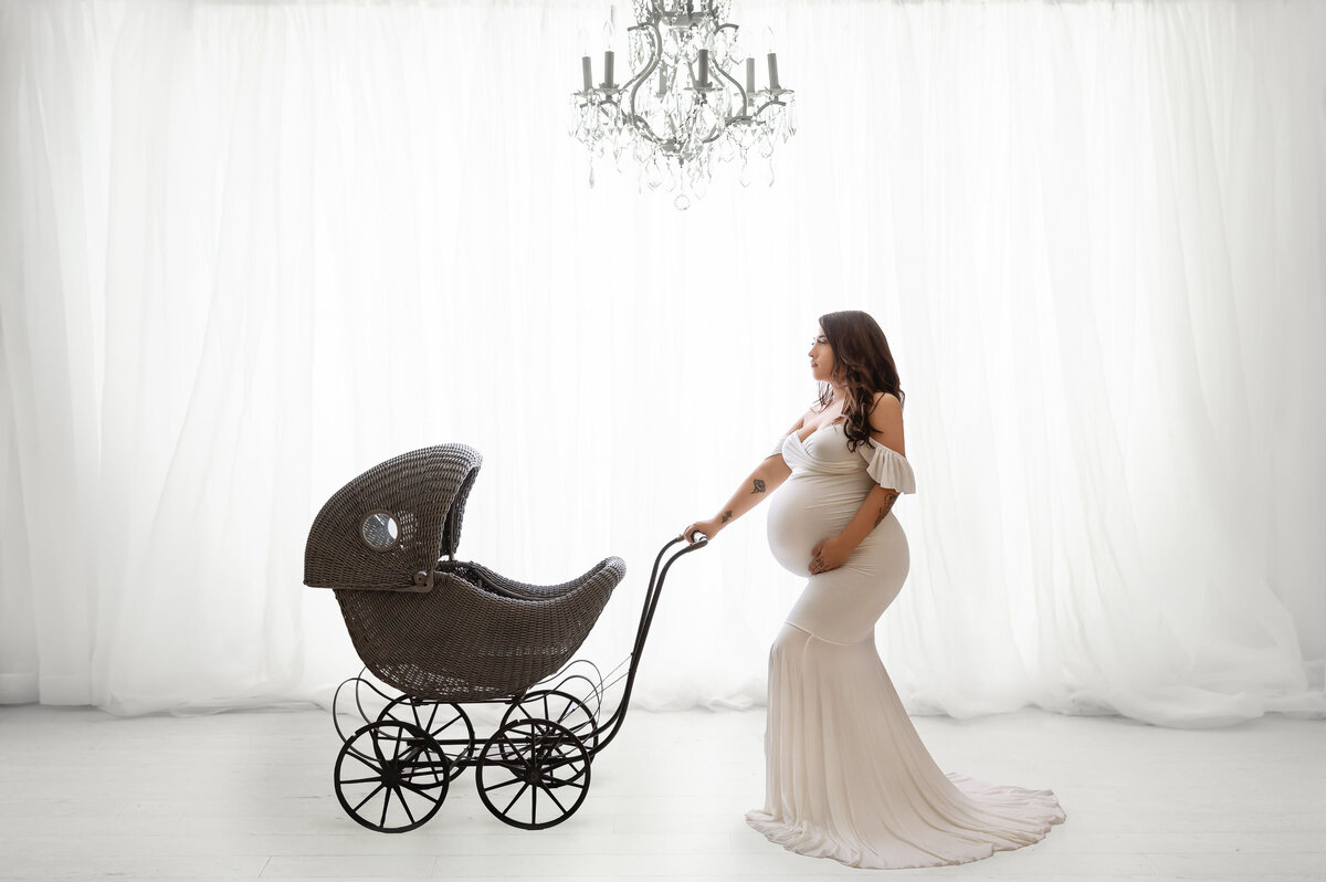 Gorgeous mother to be posed with carriage wearing a white gown in front of curtains and chandelier above with a white aesthetic  near dupont