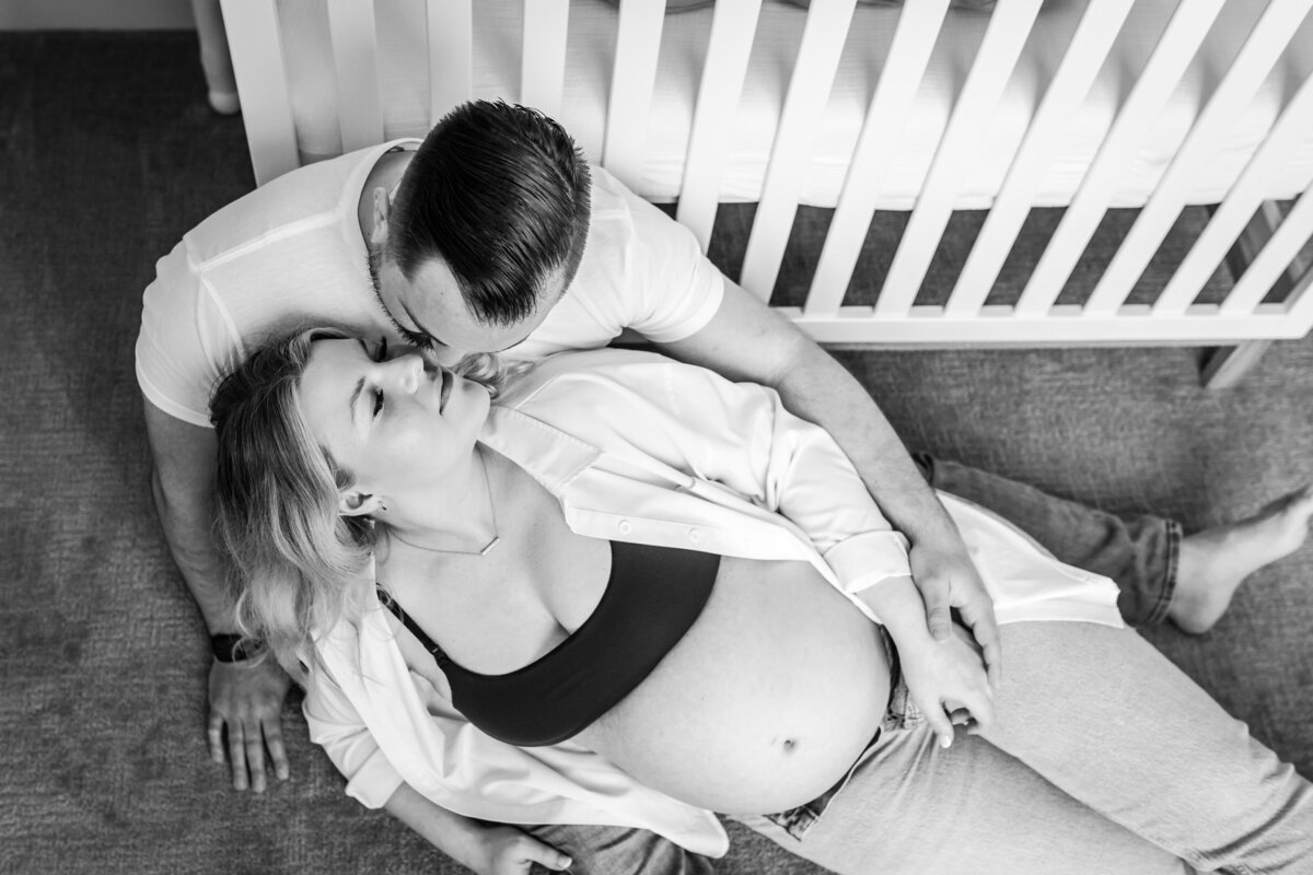 The couple sits on the floor in front of a crib, the man kissing the woman's forehead as she leans back into his arms, her baby bump prominently displayed.