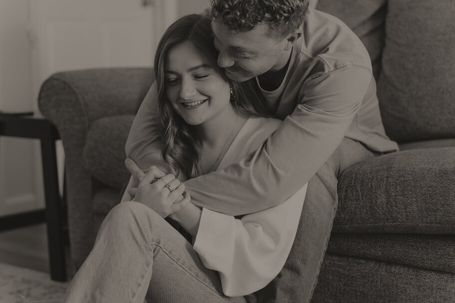 A man is sitting on a couch with his girlfriend sitting between his legs and he holds her around her shoulders. She is smiling with her eyes closed.