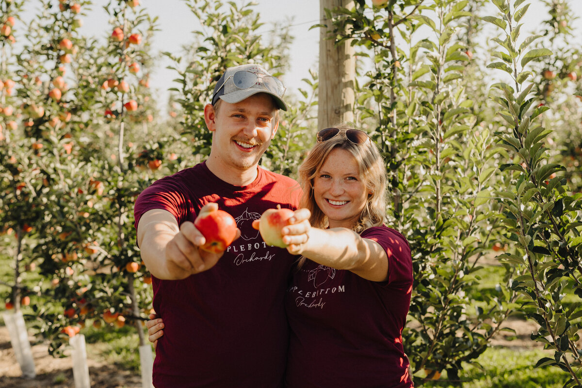 Owners, Jeff and Chelsea, each holding an apple in front of orchard while sporting Applebottom Orchard shirts.