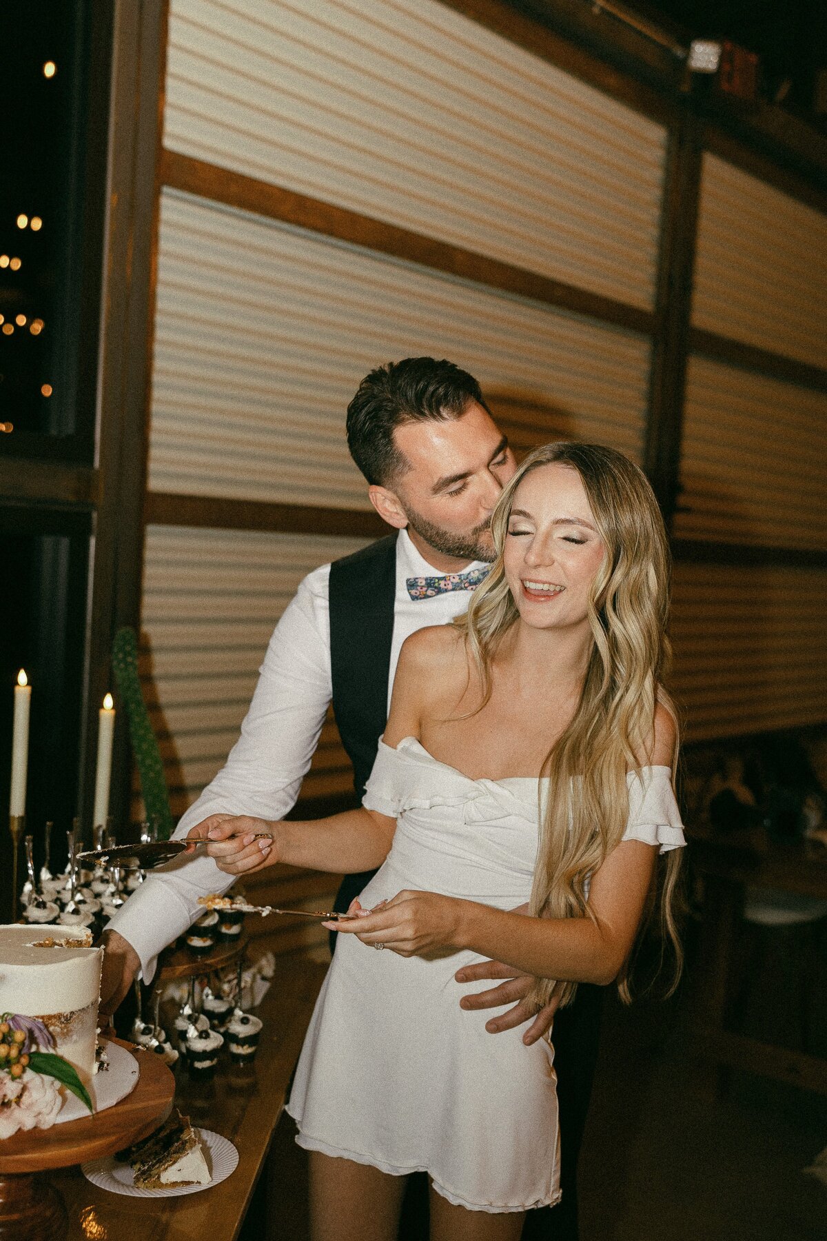 A groom kisses his bride while cutting their cake at their wedding