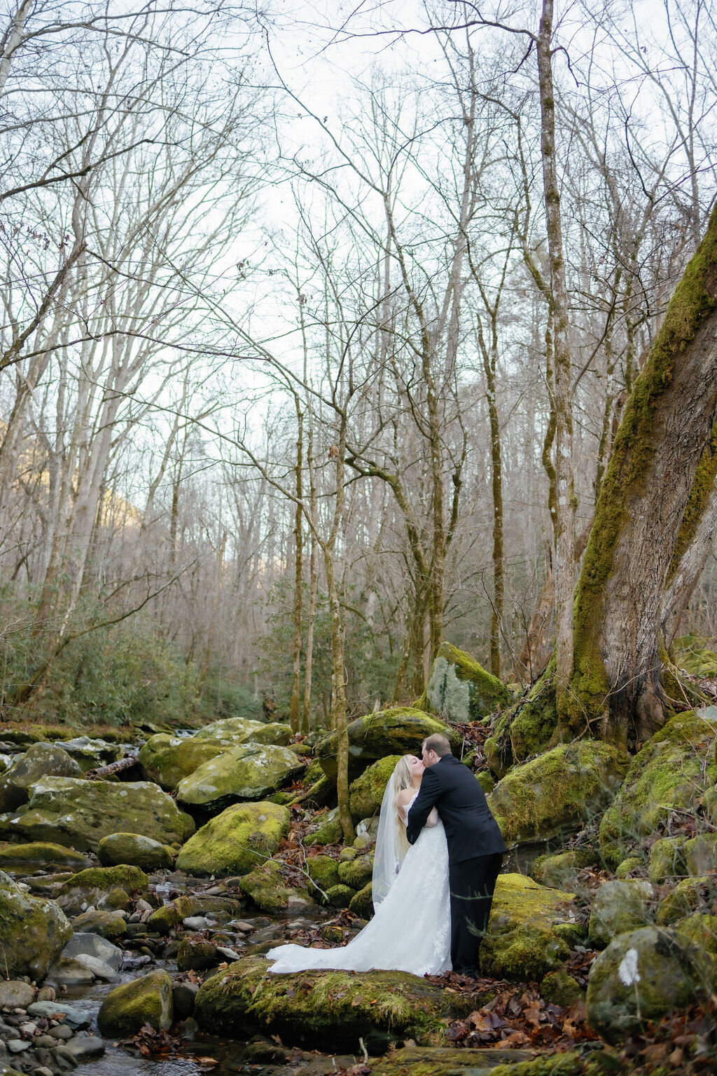 couple eloping to Gatlinburg kisses in the river surrounded by mossy rocks and bare trees for a winter wedding in the Smoky Mountains 
