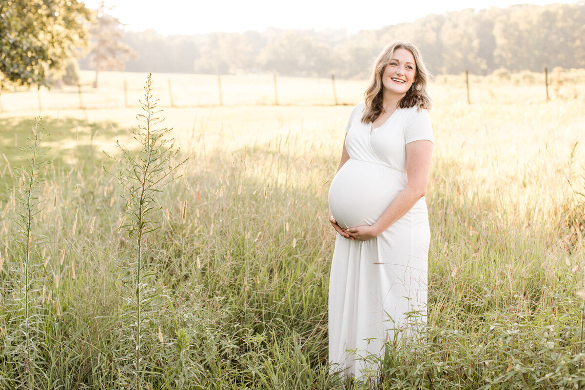 Pregnant mom stands in a field of tall grass holding bump and smiling off camera during maternity session in Lawrenceville at Prospect Farms