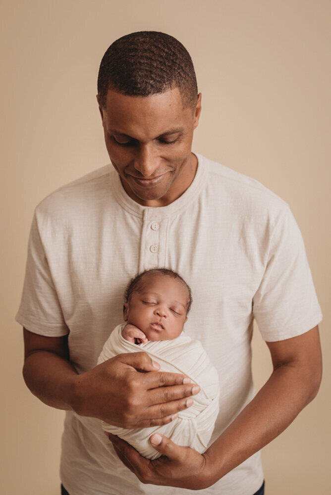 Newborn photos with Atlanta, Georgia newborn photographer of sleeping baby boy with hands under chin wrapped in cream swaddle being held by his dad.