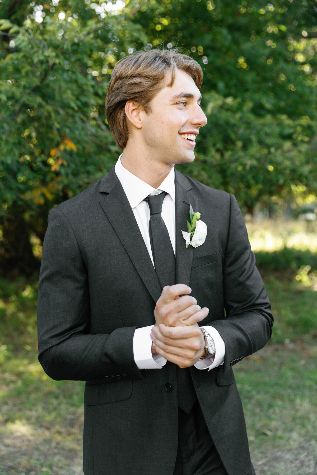 Groom in classic black suit with white boutonnière standing outdoors at an Arkansas wedding, featuring simple and elegant floral pin design.