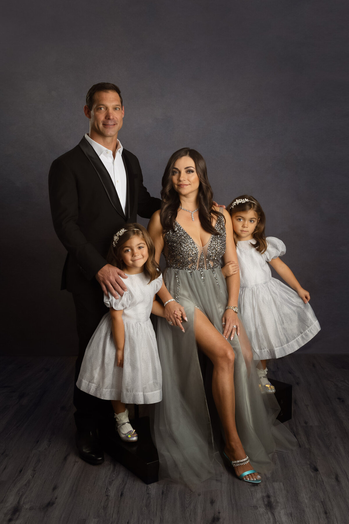 formal studio portrait of a family of 4. they are seated and wearing formal evening wear. backdrop is dark grey