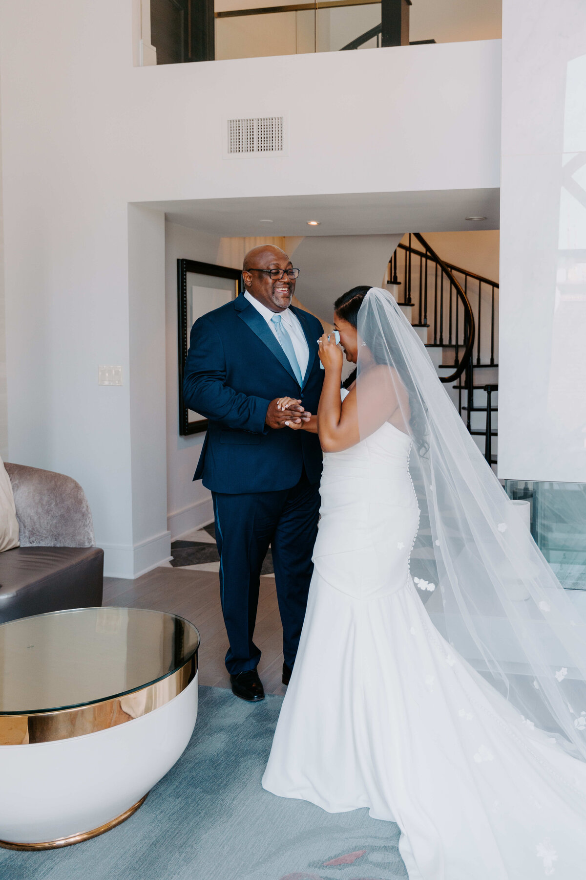 Bride in a white gown and veil shares an emotional first look moment with her father indoors before the wedding ceremony