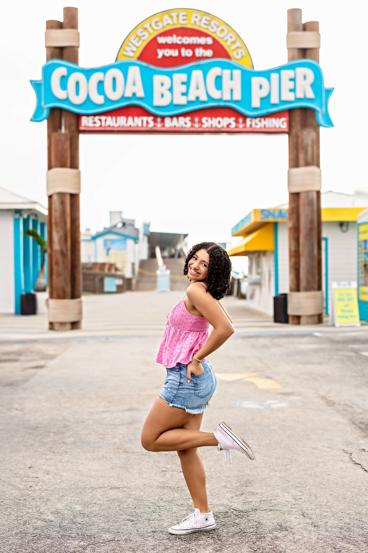 Senior girl posting confidently beneath the iconic Cocoa Beach Pier sign, one leg kicked up in jean shorts and a pink top, playful and bold.