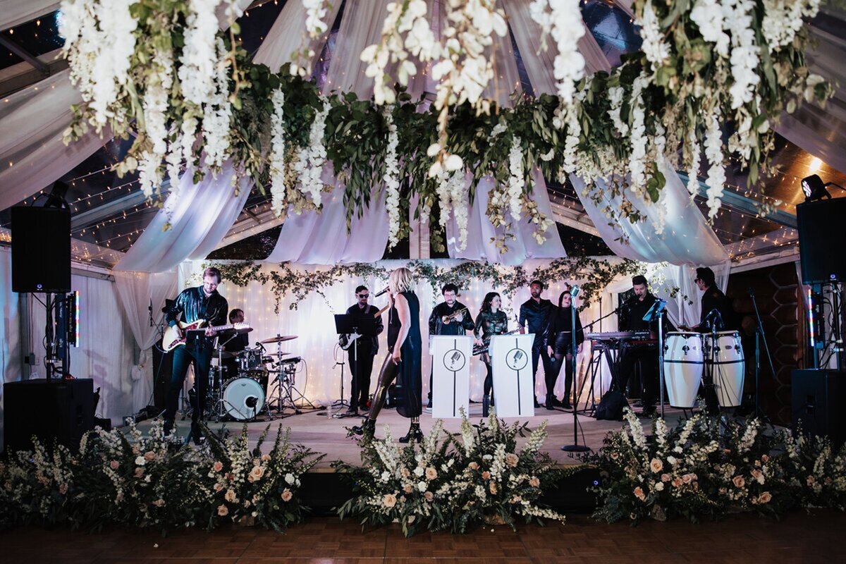 A band is set up on stage in a wedding tent, a chandelier drips flowers overhead