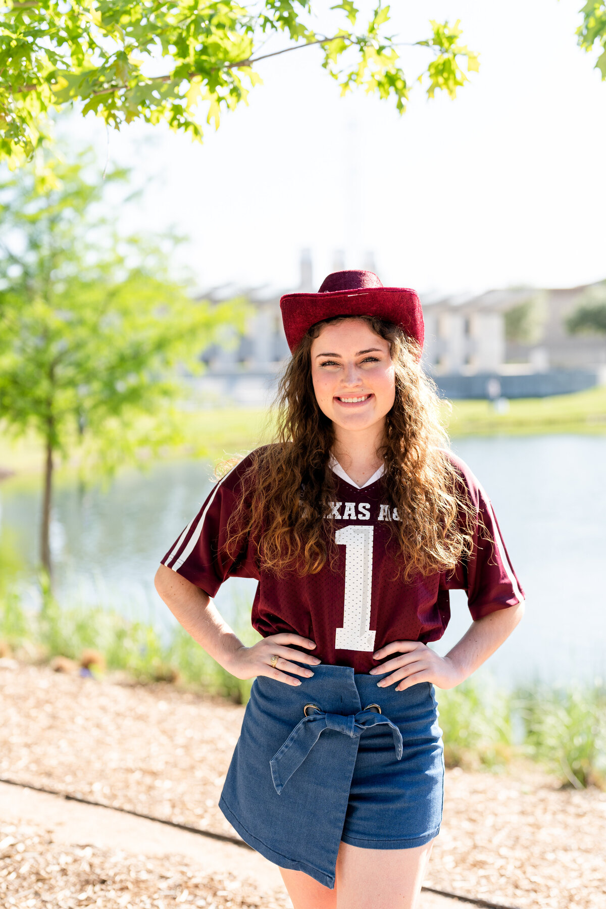 Texas A&M senior girl with hands on hips smiling wearing maroon jersey and cowboy hat and jean skirt in Aggie Park