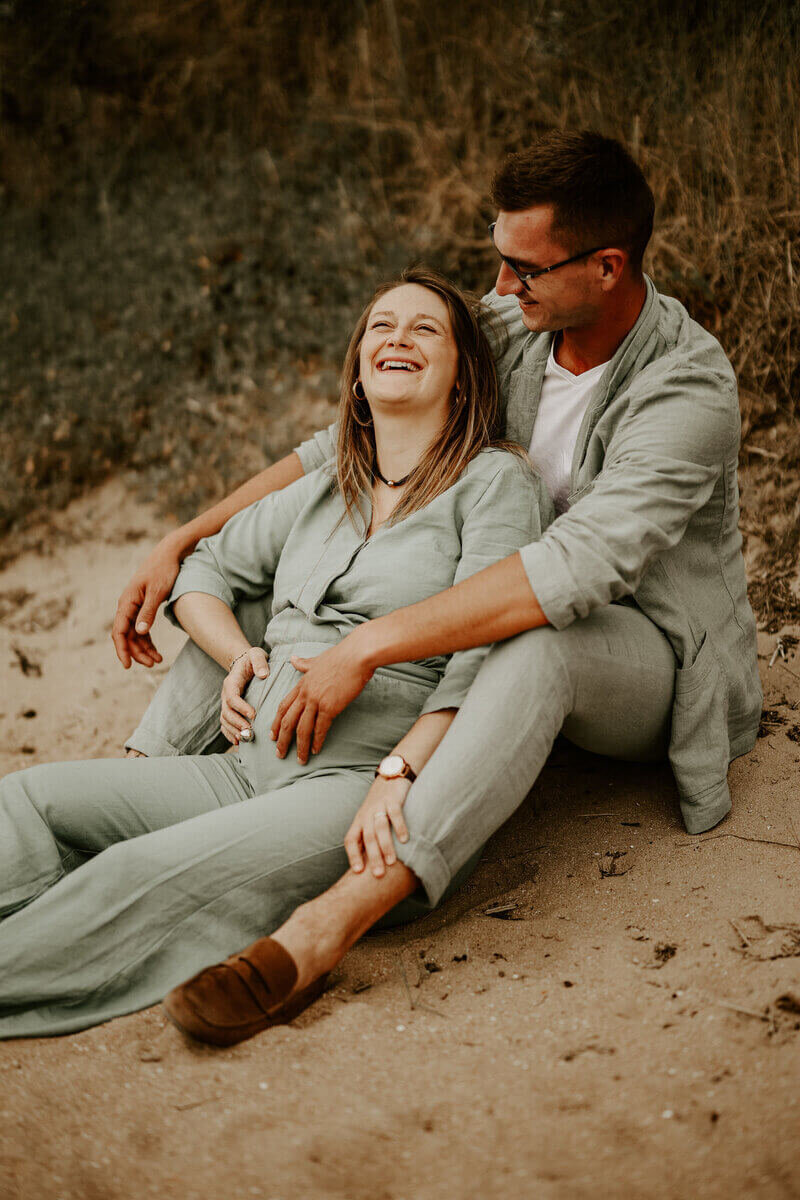 Femme enceinte, souriant, assise entre les jambes de son homme la regardant, dans une décor de dune. Photo prise par Laura, photographe grossesse en vendée.