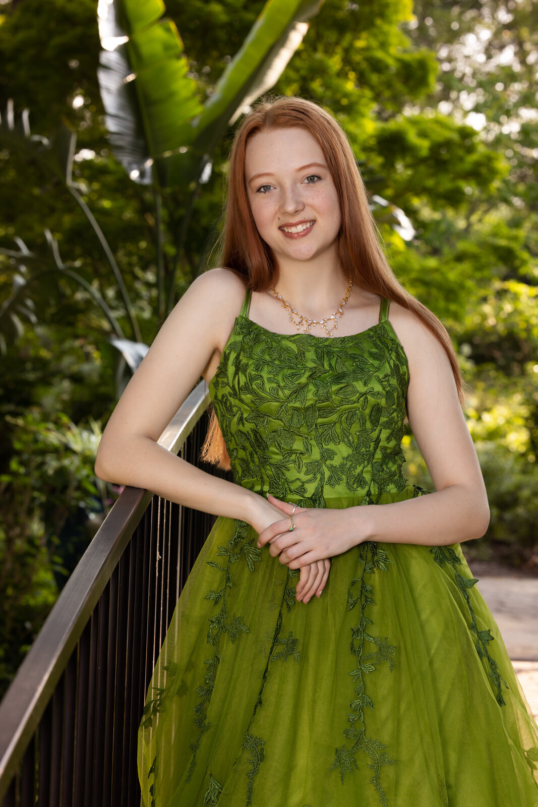 senior-girl-in-green-dress-leaning-on-fence-with-greenery-surround