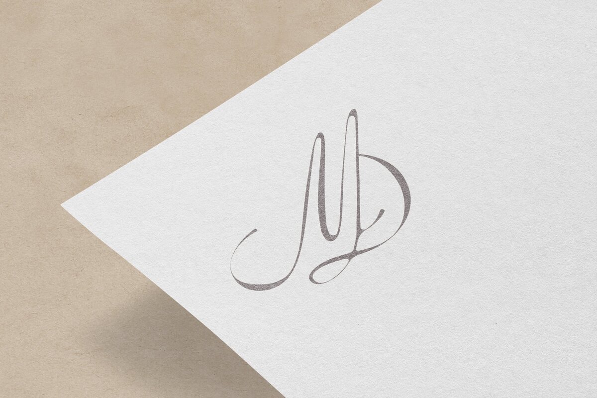 A corner of a white piece of paper displays a stylized, elegant monogram with the initials "MD" in gray cursive font on a beige background, showcasing the meticulous work of an expert brand designer.