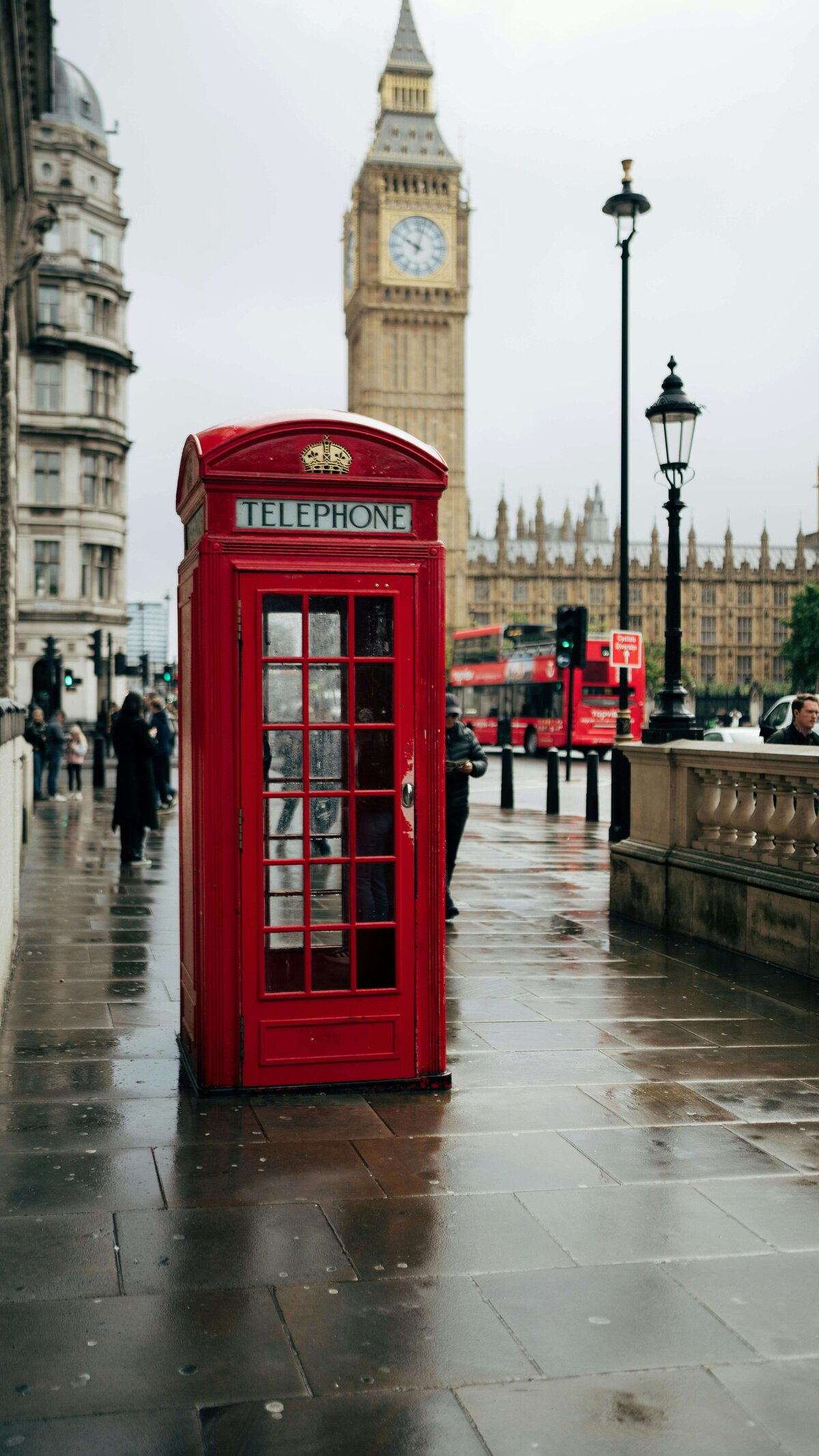 A classic red telephone booth stands on a rainy street in London, with Big Ben and a double-decker bus visible in the background. This quintessential British scene captures the timeless charm of travel experiences enabled through vacation fundraising for charities.