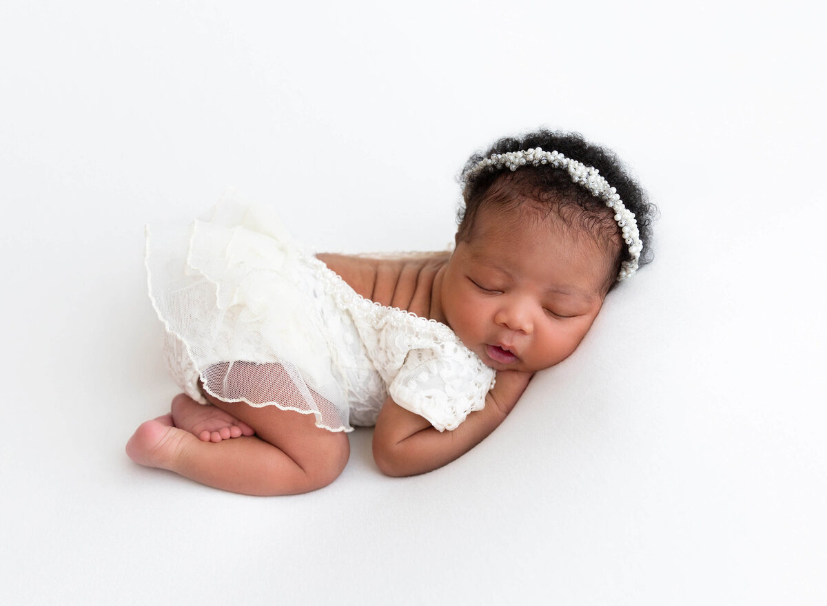 Baby girl in a one lace onesie is sleeping on her belly. Image is shot side profile. Her hands are under her chin and her face is turned toward the camera. Image taken by Rochel Konik Photography.