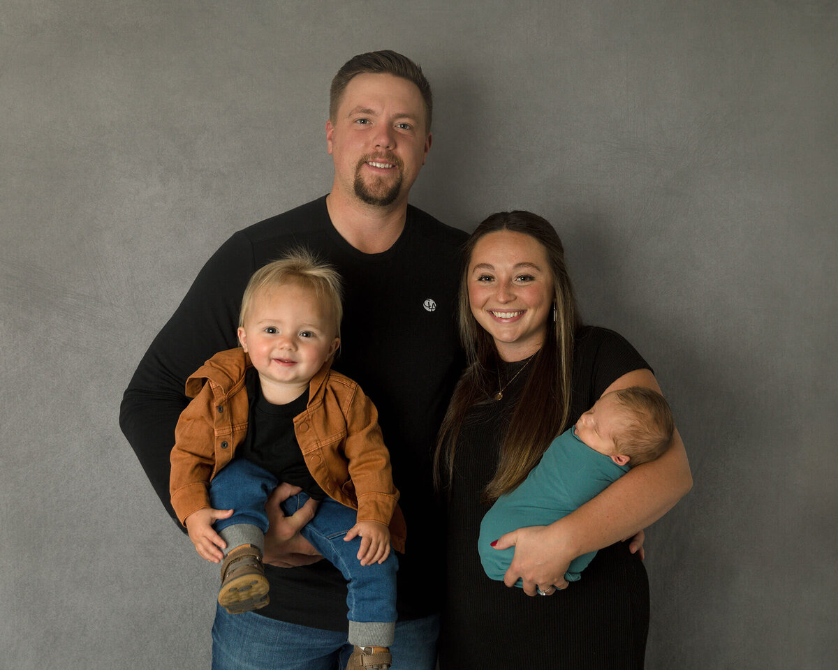 Dad and Mom holding toddler boy and newborn boy in front of gray background.