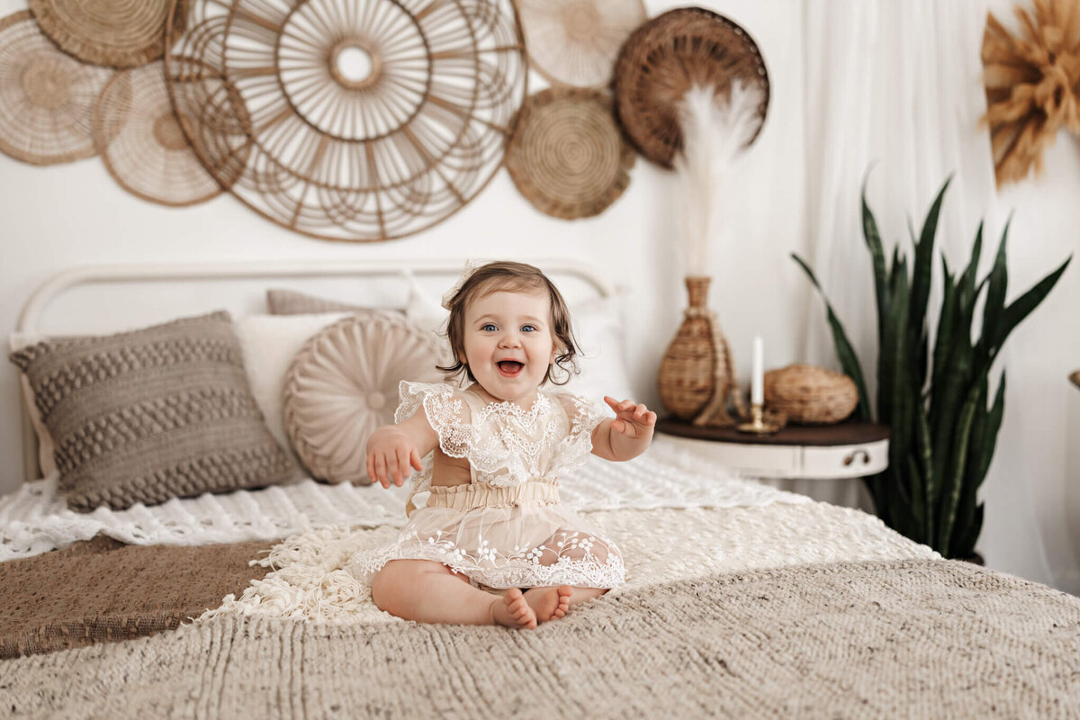A smiling baby in a lace outfit sits on a bed with textured blankets, captured beautifully by a portrait photographer. The background, featuring woven wall decor, pillows, and plants, creates a cozy bohemian atmosphere perfect for family photography in Bloomington, Illinois.