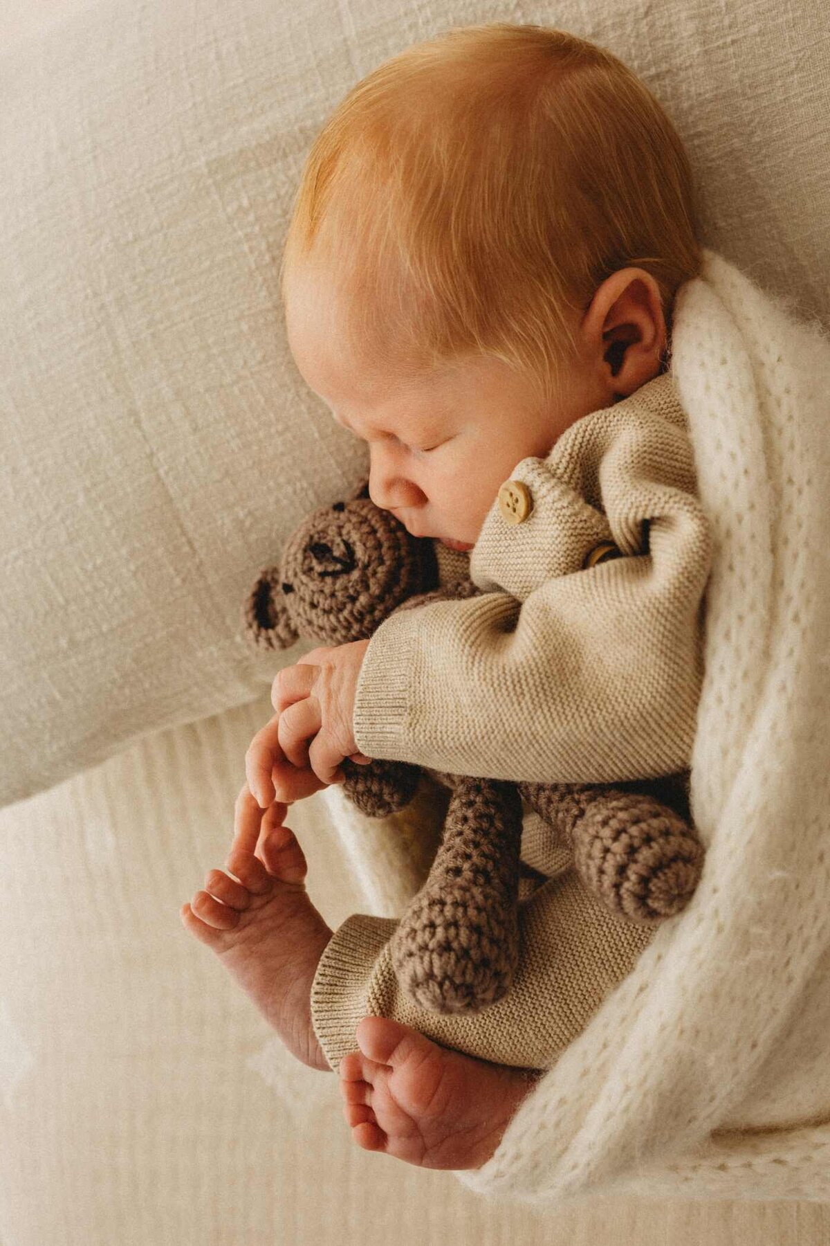 Ruua Hamdan Photography (UK): A newborn baby with red hair sleeps peacefully, cuddling a small brown teddy bear, wrapped in a soft white blanket.