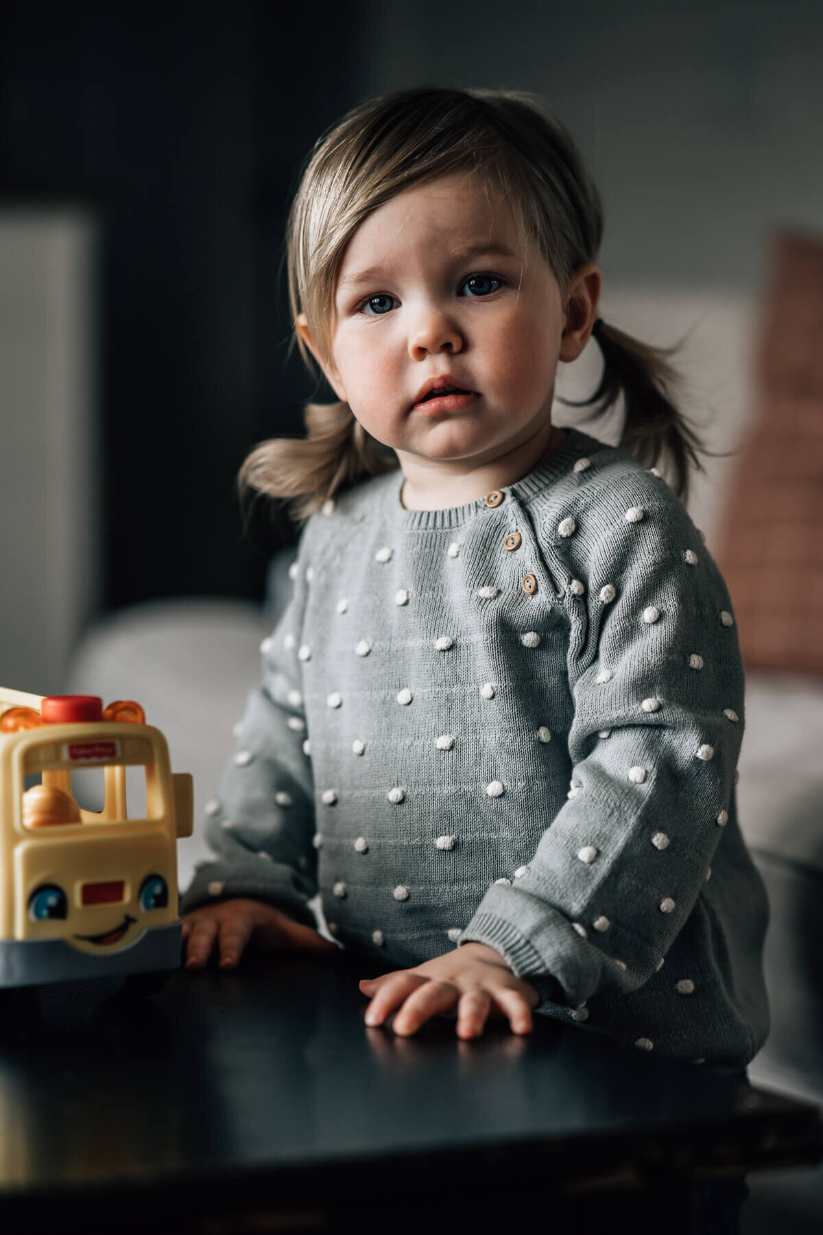 A two year old girl with pigtails and side swept bangs stands at her living room coffee table during an in home photo session by Kate Simpson in Minneapolis.