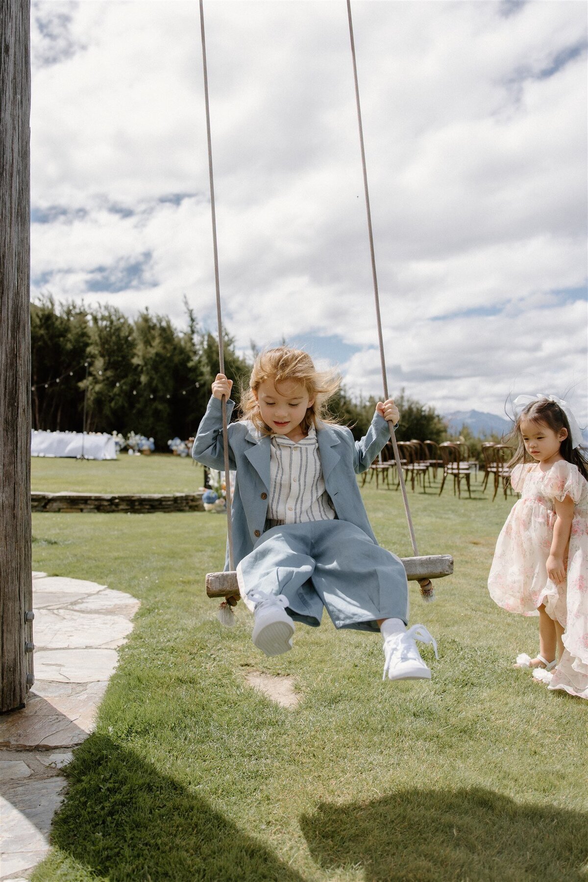 Young boy in a pale blue pantsuit happily swinging on a wooden swing while a girl in a floral dress watches, with wedding chairs and trees in the background.