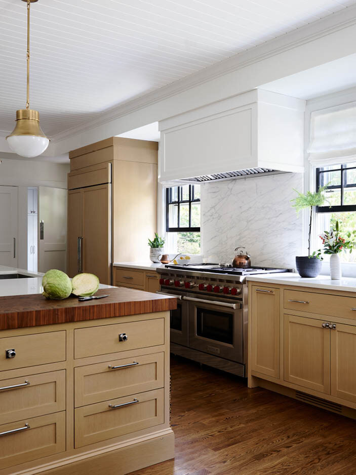 View the kitchen in a historic Hingham home, featuring a custom hood integrated with the soffit, a slab marble backsplash, and a range perfectly framed by original windows.