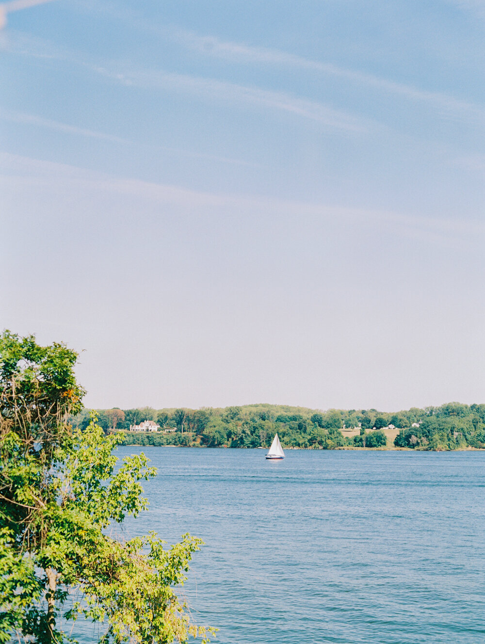 The serene scene of a sailboat on a calm, expansive lake under a clear blue sky was captured at The Hamptons, New York, waterfront estate during a tented wedding by photographer Bonnie Sen. Green foliage and trees elegantly frame the left side of the image, and one can see more trees and land in the background across the peaceful water. East West Films also provided videography services for the event.