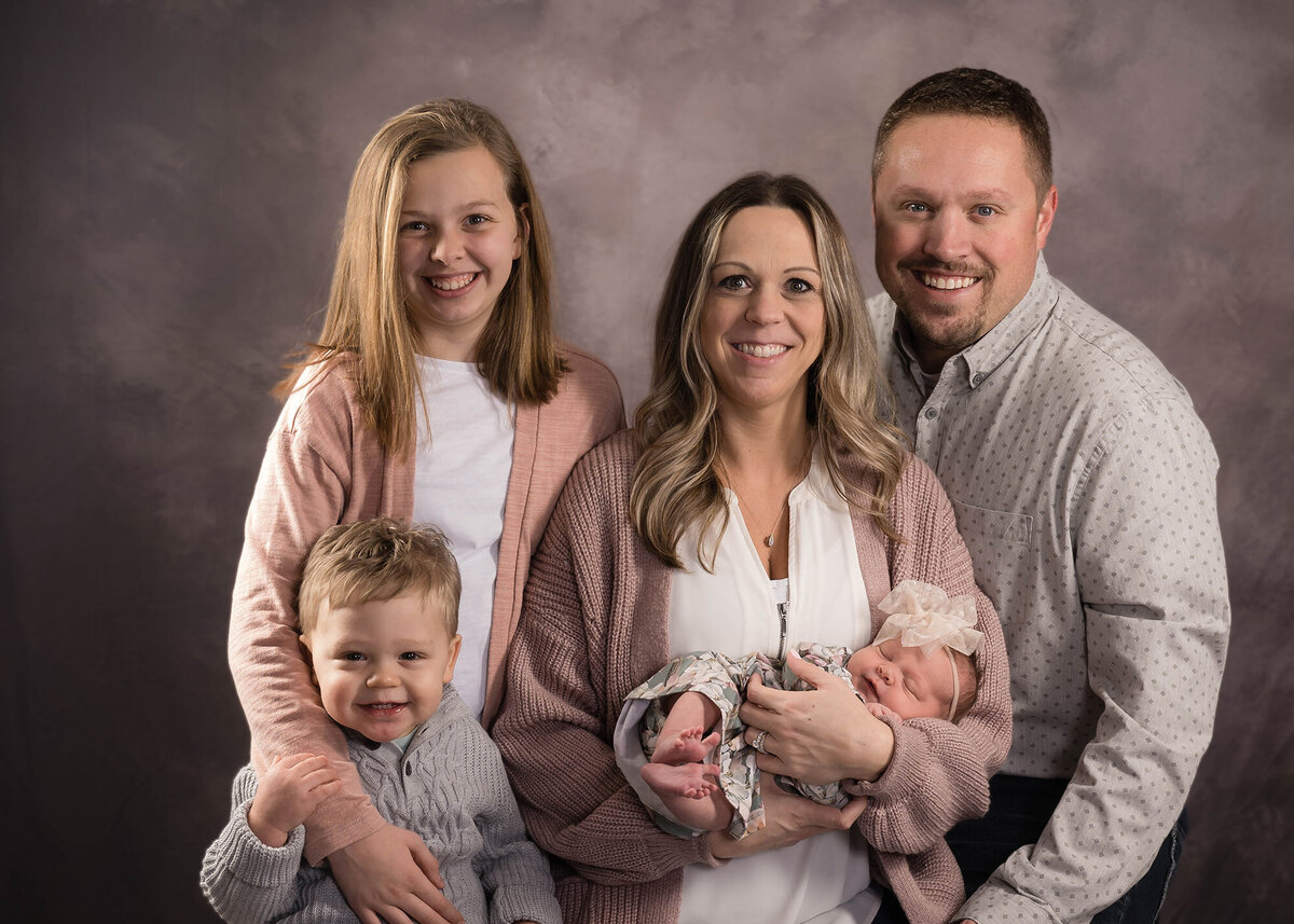 A family of 5 stands in a studio with a newborn in mom's arms from an Iowa Newborn Photographer