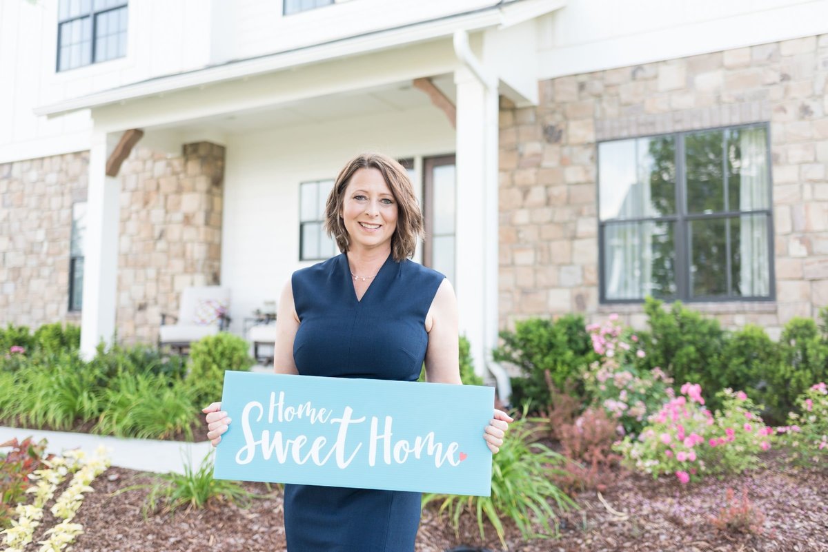 Realtor in a navy dress holding a blue sign that says "home sweet home" outside of a Nashville home