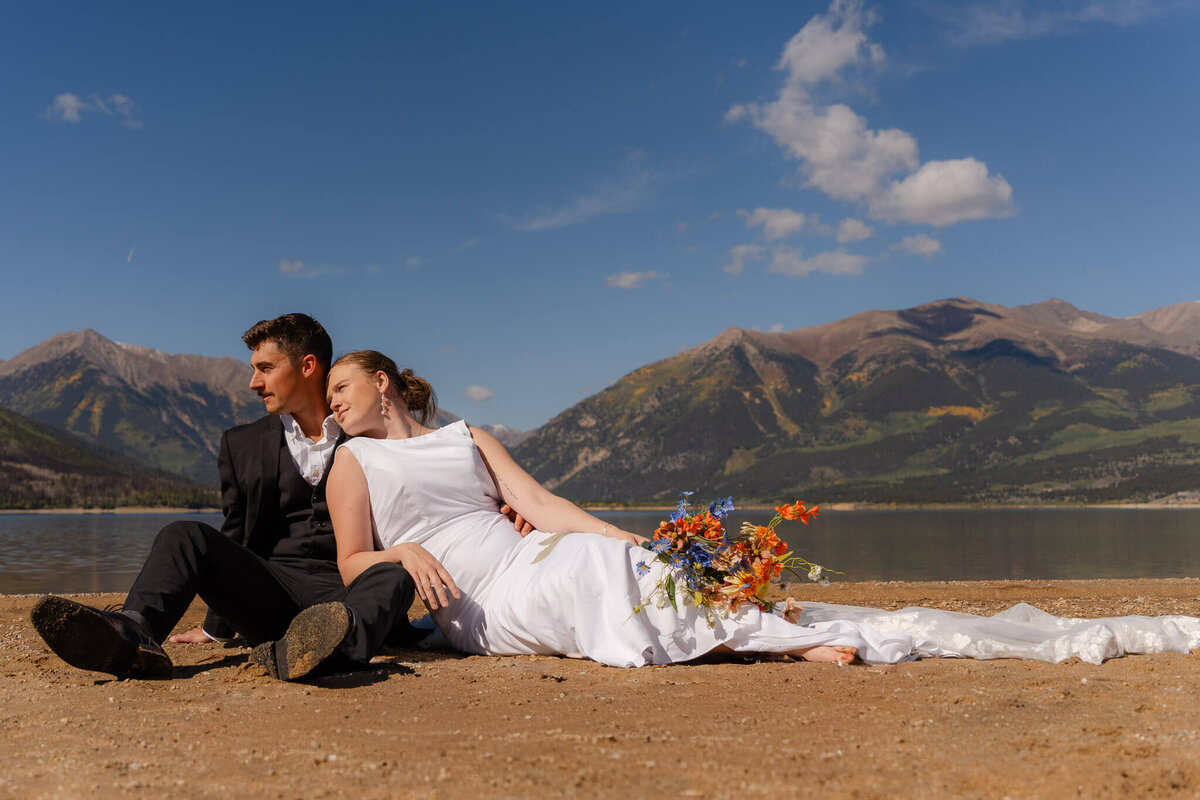 bride and groom sitting together on the beach of an alpine lake in Twin Lakes Colorado with the mountains in the background