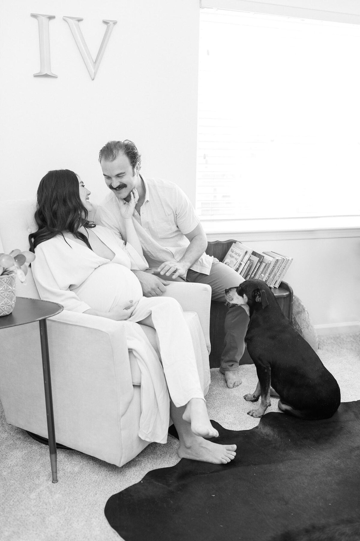 Virginia Beach Maternity Photography of a pregnant woman and her husband sitting in the nursery with their dog, smiling at each other in black and white by Mary Eleanor Photography