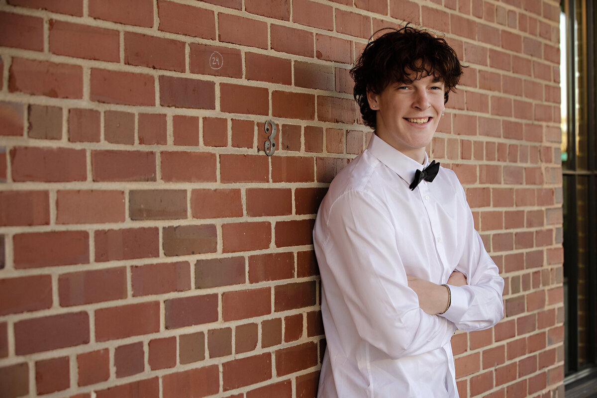 Senior portrait leaning against a brick wall in a formal casual outfit with bowtie and spring textures, photographed in Charlotte NC.