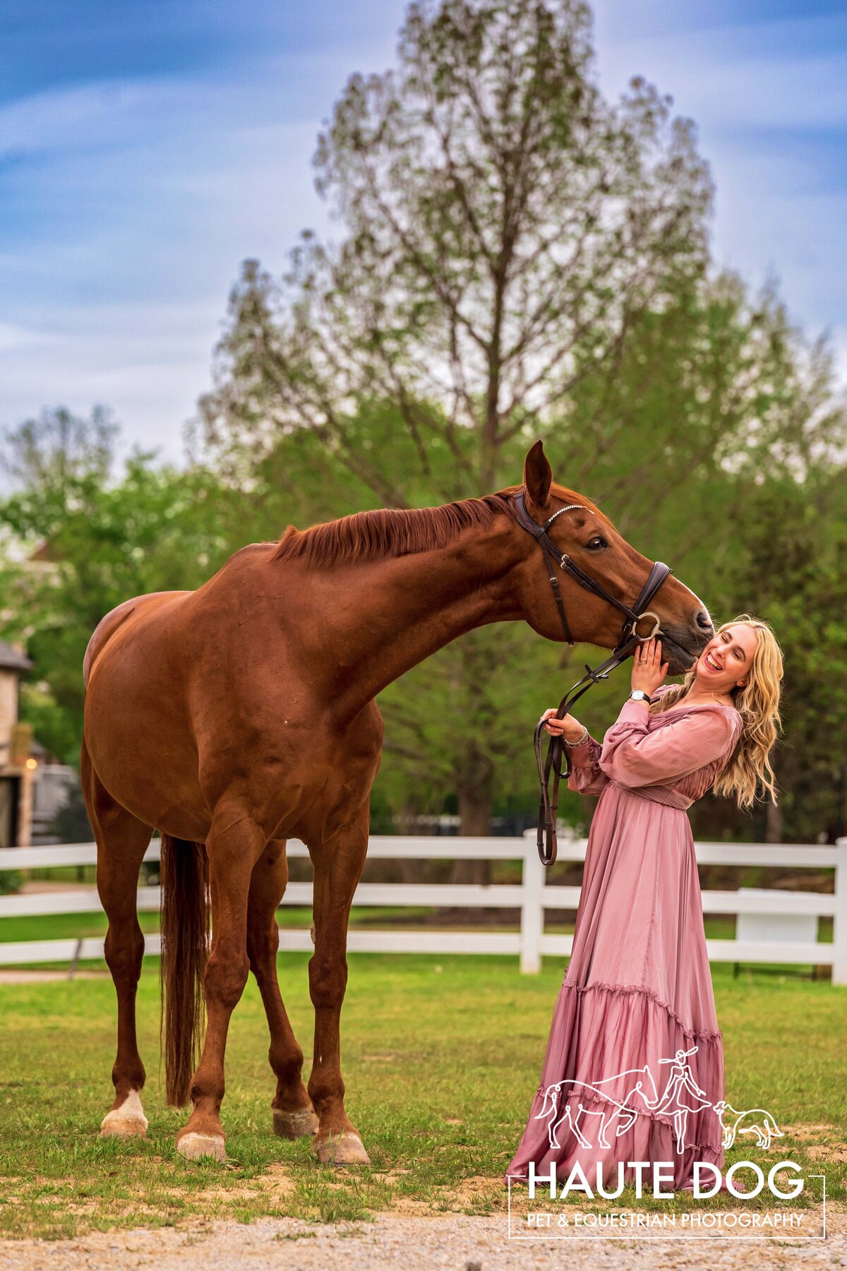 A tall chestnut horse kisses equestrian wearing a long pink dress on the cheek while she laughs.