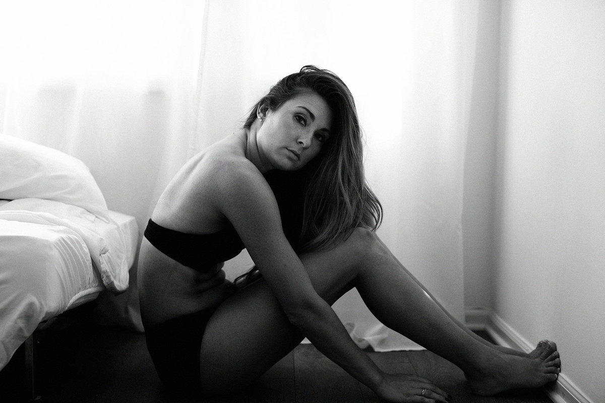 black and white portrait of a woman in black lingerie leaning next to a bed