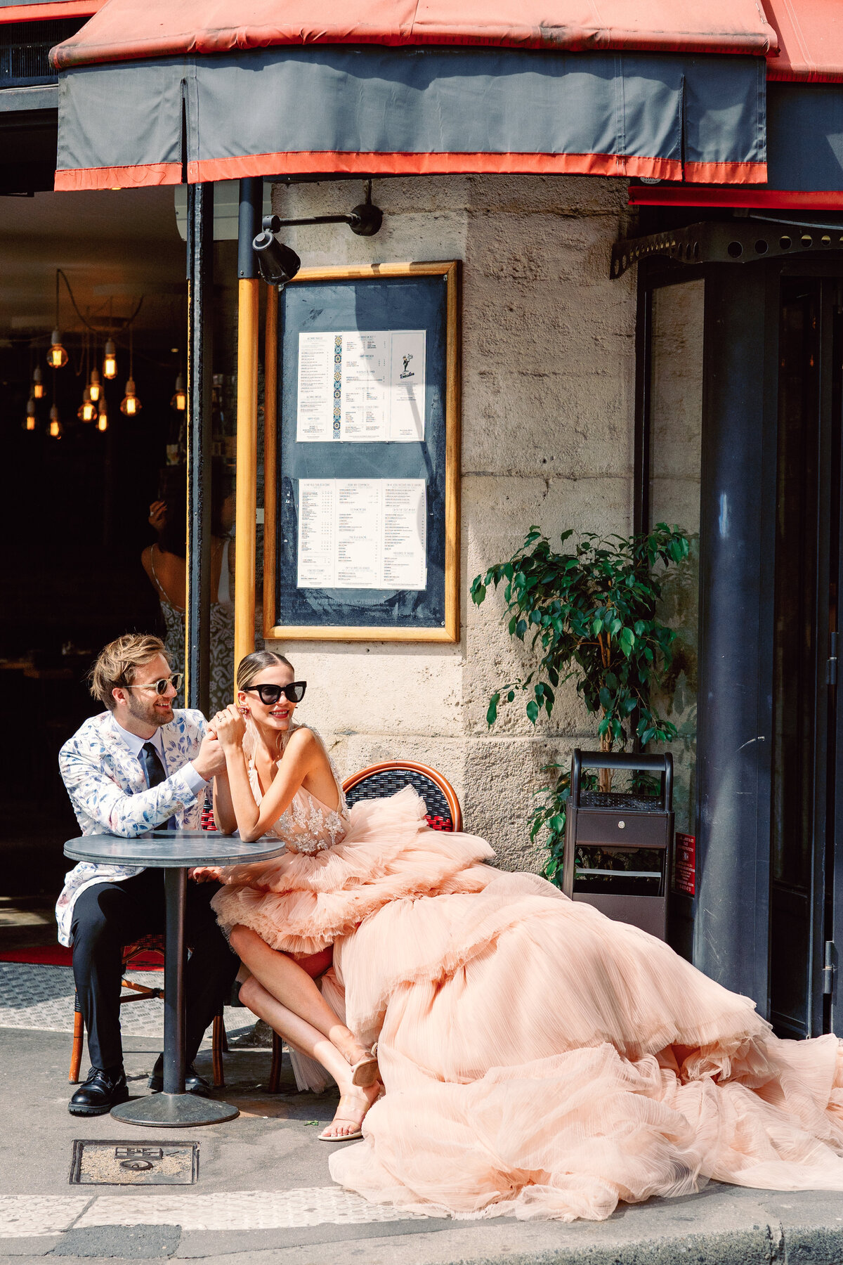 Couple at a cafe enjoying champagne, she in a peach gown with long train at their Paris engagement session, photographed by Italy wedding photographer.